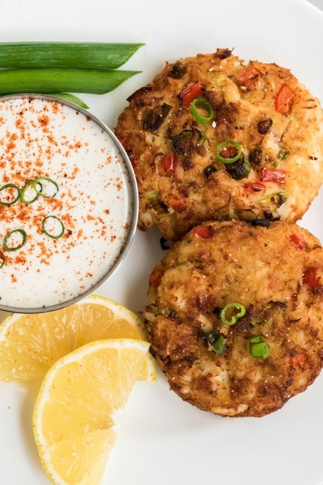 Dois bolos de caranguejo de fritadeira em um prato branco com molho e fatias de limão