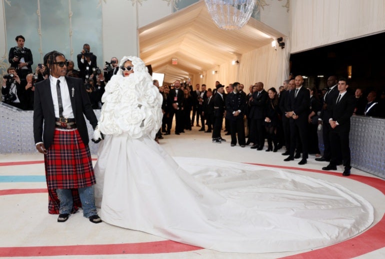 Rihanna e seu parceiro ASAP Rocky posam para fotógrafos no Met Gala.  Ela está usando um vestido branco com uma longa cauda.  Ele está em um kilt xadrez com camisa branca e jaqueta preta.