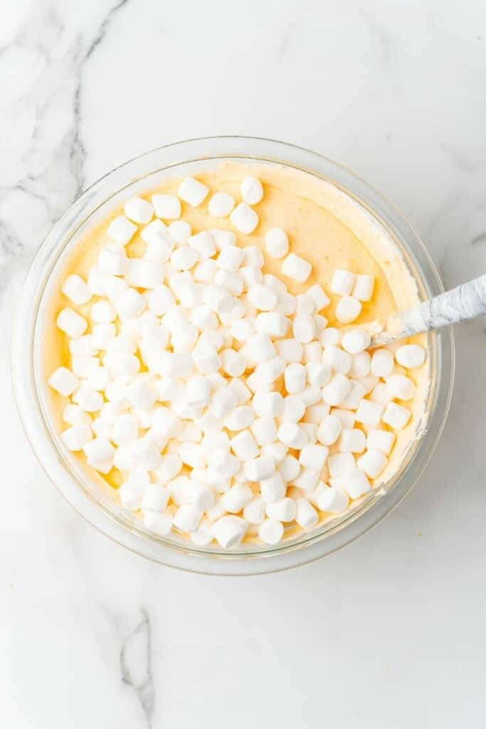Pequenos marshmallows em cima de Orange Dreamsicle Salad em uma tigela transparente com uma colher.