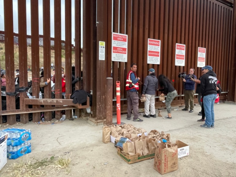 Voluntários entregam comida e água pelas aberturas em uma parede de metal