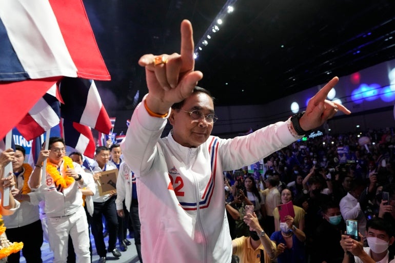 Prayuth gesticulando para seus apoiadores.  Ele está vestindo um agasalho nas cores da festa, vermelho, branco e azul, e há bandeiras ao fundo