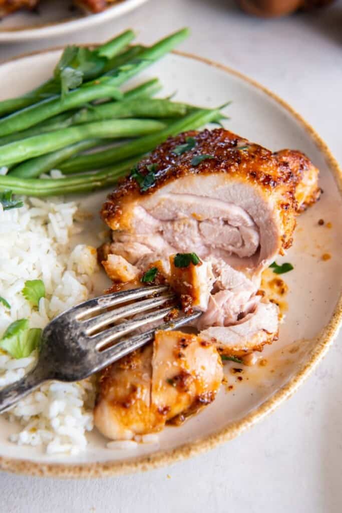 Um garfo segurando um pedaço de coxa de frango desossada em um prato branco com arroz e feijão verde.