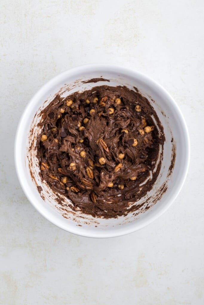 Dobrar nozes tostadas, pedaços de caramelo e lascas de chocolate com a massa de biscoito em uma tigela branca.