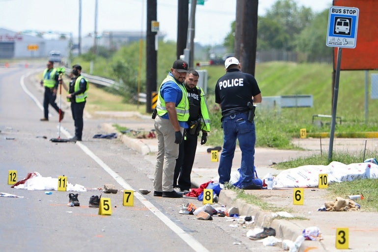 A polícia investiga o local do incidente.  Eles marcaram itens na estrada com números.  Há uma placa de ponto de ônibus ao lado da estrada.