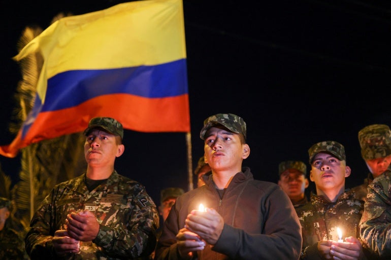 Soldados contra um céu noturno escuro, vestidos com uniformes militares, seguram velas e uma grande bandeira da Colômbia.