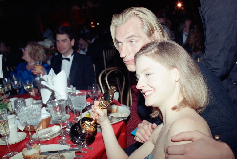 Jodie Foster, com um sorriso no rosto e seu Oscar na mão, senta-se com Julian Sands durante o Baile do Governador no Shrine Auditorium em 29 de março de 1989. Sands a abraça.