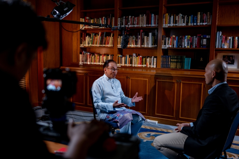 Anwar Ibrahim falando para 101 East.  Ele está vestindo uma roupa tradicional malaia.  Suas mãos estão abertas enquanto ele explica um ponto.