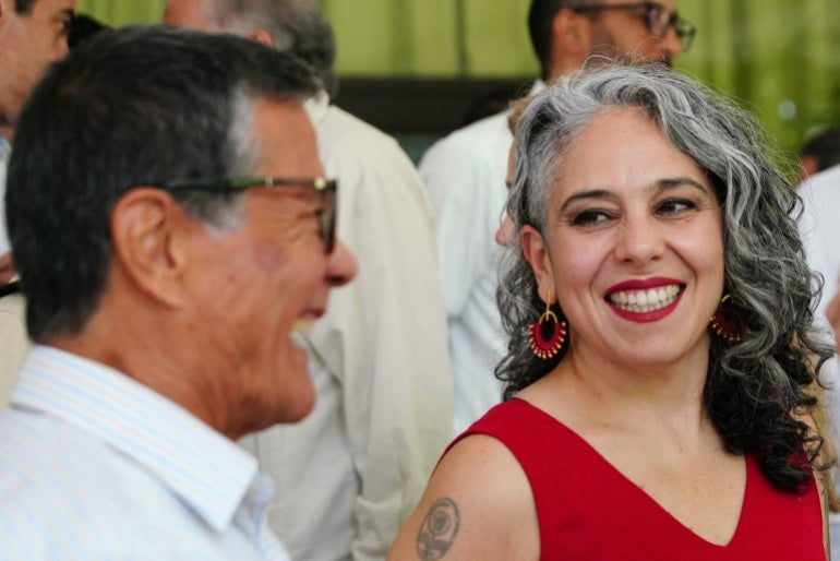 Uma mulher de vestido vermelho sorri e olha para um homem de camisa branca e óculos.