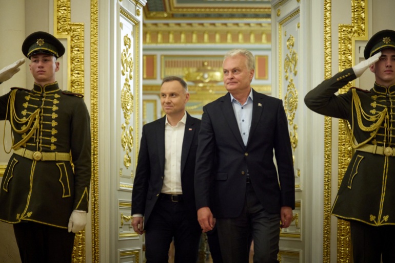 O presidente da Polônia, Andrzej Duda, e o presidente da Lituânia, Gitanas Nauseda, chegam para uma coletiva de imprensa conjunta com o presidente da Ucrânia, Volodymyr Zelenskyy.  A sala é ornamentada e há dois soldados em trajes cerimoniais de cada lado da porta.