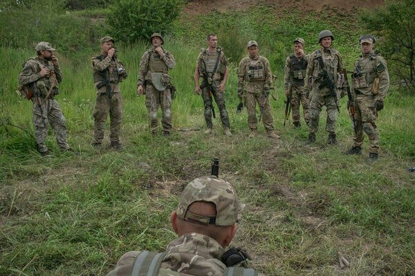 Soldiers in camouflage stand in front of another soldier in a green field.