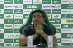 Abel Ferreira gesticulando em coletiva de imprensa, com microfones e logo do Palmeiras visíveis.