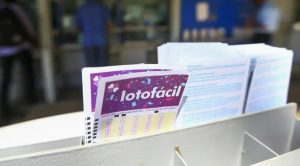 Mãos segurando bilhete da Lotofácil e notas de dinheiro, simbolizando o prêmio milionário.