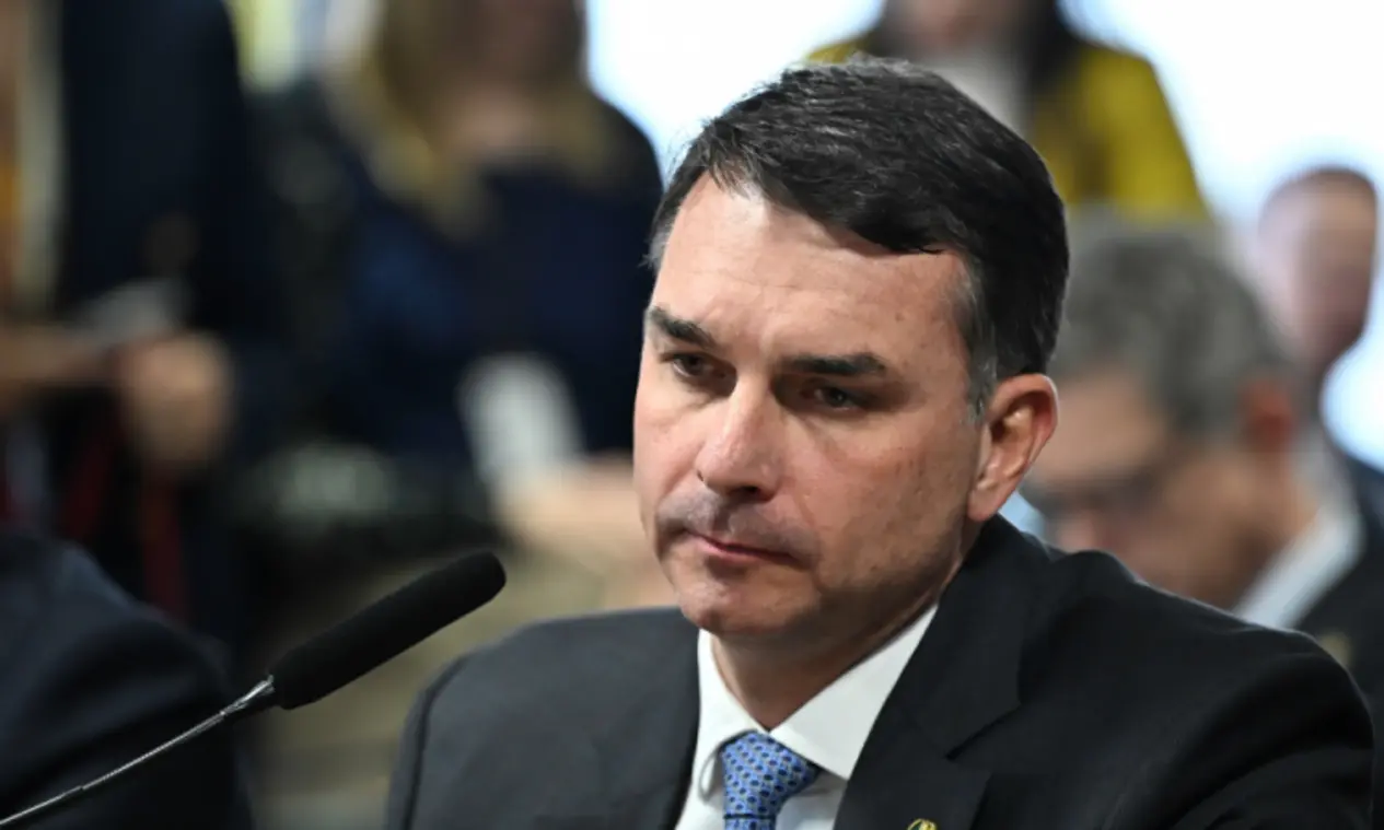 Foto do senador Flávio Bolsonaro, vestindo terno, discursando em um púlpito durante o evento CPAC no Texas, com fundo desfocado.